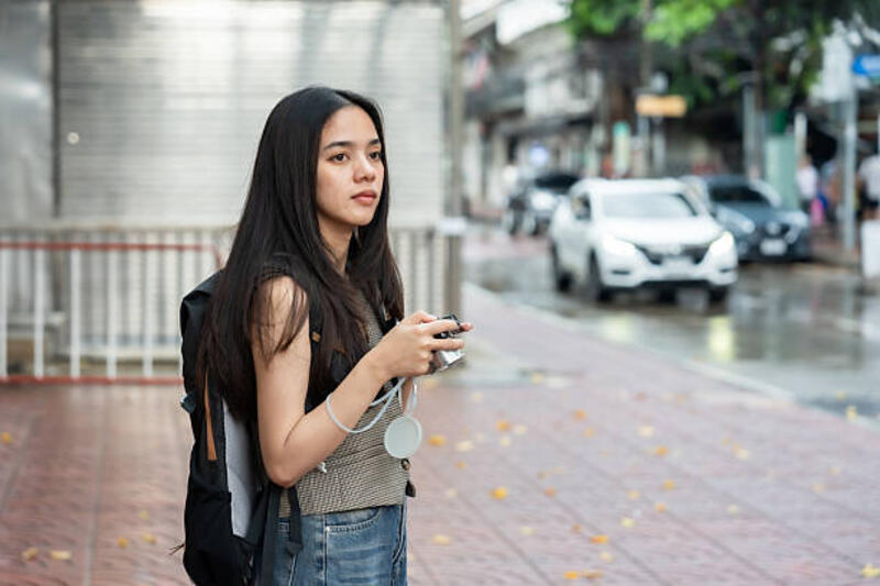 Beautiful Asian solo traveler standing on a wet city sidewalk holding a vintage camera. Young female tourist in casual attire and backpack looking around, moments of Bangkok