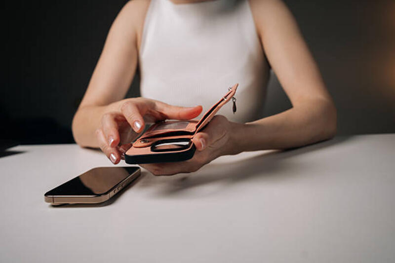 Woman sitting at white table holding new salmon-colored wallet-style phone case with magnetic closure while old phone lies on table. Woman sitting at white table holding new salmon-colored wallet-style phone case with magnetic closure while old phone lies on table. Concept of modern technology and stylish accessories. Magnetic Phone Wallet stock pictures, royalty-free photos & images