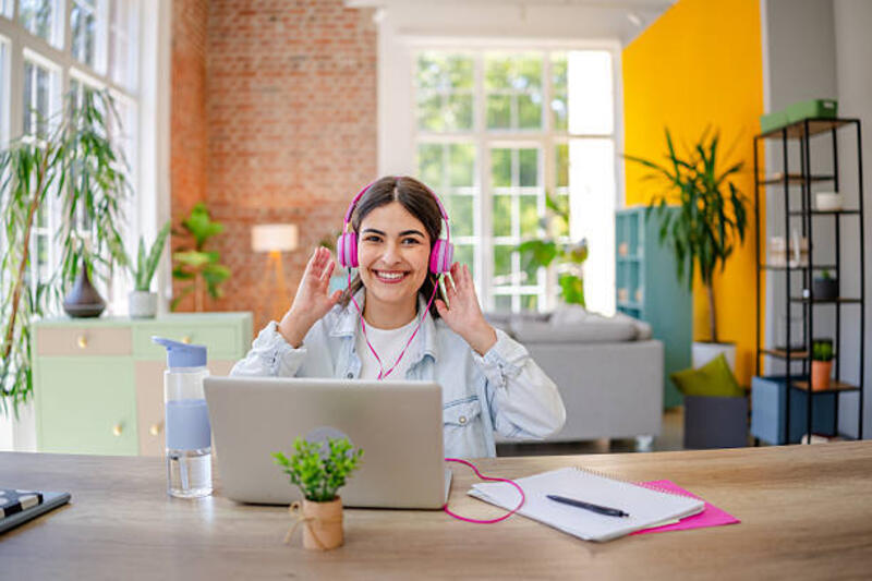 Young woman enjoying her time at home while working on a laptop in a bright and cheerful living space A young woman is smiling and wearing headphones while sitting at her desk in a lively living room. She engages happily with her laptop, surrounded by plants and colorful decor. listening to music stock pictures, royalty-free photos &amp; images