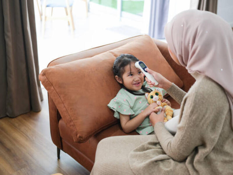 a woman using a digital thermometer to check the temperature of a young girl holding a toy on a couch in a home setting - termometer digital potret stok, foto, &amp; gambar bebas royalti