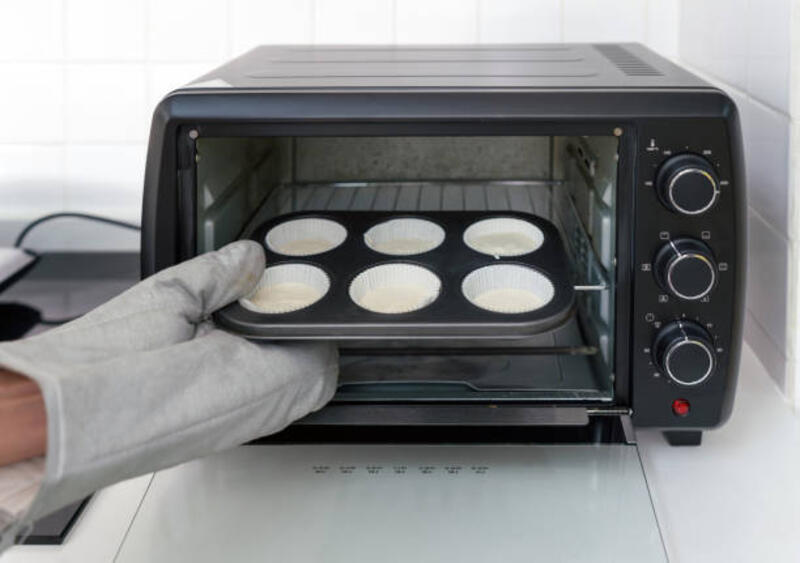 Tangan dengan sarung tangan oven, letakkan nampan muffin dengan adonan ke dalam oven pemanggang roti hitam di dapur putih.