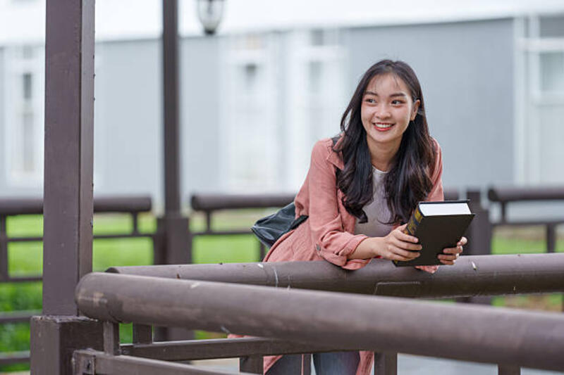 mahasiswi-wanita-muda-tersenyum-sambil-bersandar-di-pagar-memegang-buku-di-luar-ruangan-di.jpg (612×408)