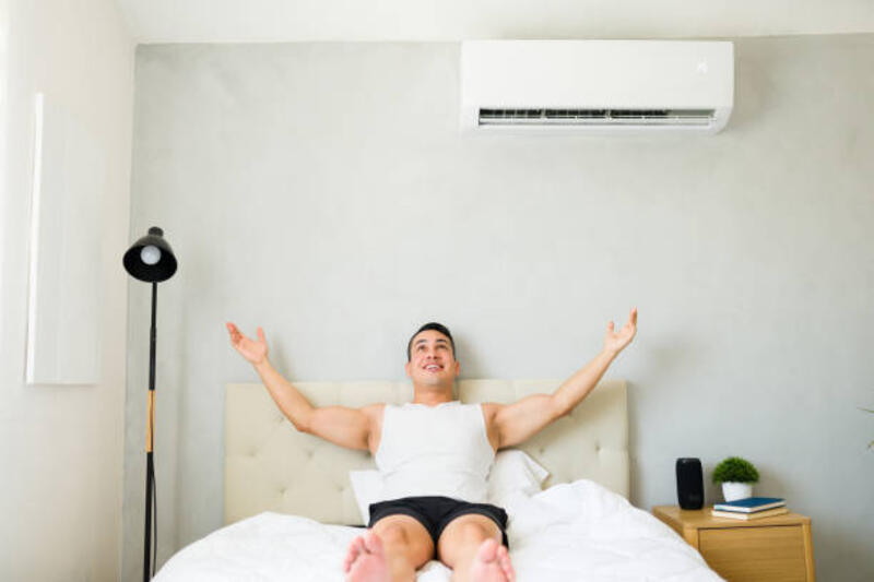 Pria latin yang ceria bersantai di tempat tidur dengan tangan terbuka, menikmati angin sejuk dari AC di kamar tidur selama kehangatan musim panas