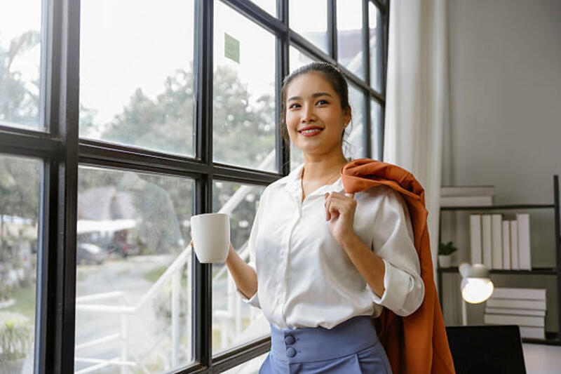 potret seorang pengusaha muda asia yang tersenyum dengan kemeja putih dan celana biru, memegang cangkir kopi dan menutupi jaketnya, berdiri di dekat jendela kantor dan melihat kamera - kemeja kemeja dan jaket wanita potret stok, foto, &amp; gambar bebas royalti
