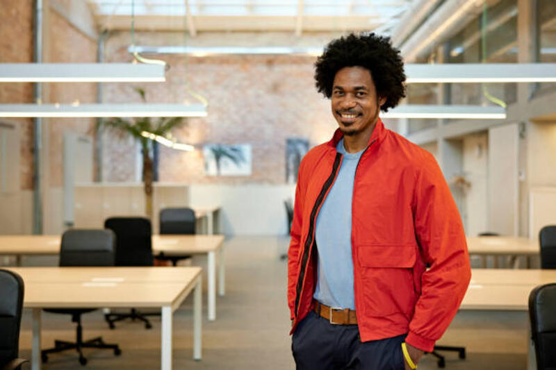 Portrait of Cheerful Black Businessman in Casual Attire Waist-up view of man in late 30s standing in industrial-style modern office with hands in pockets and smiling at camera. windbreaker jacket stock pictures, royalty-free photos &amp; images