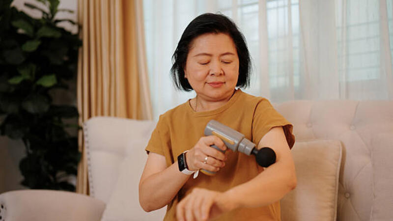 Wanita Asia dewasa memijat lengannya dengan pistol pijat di sofa di rumah