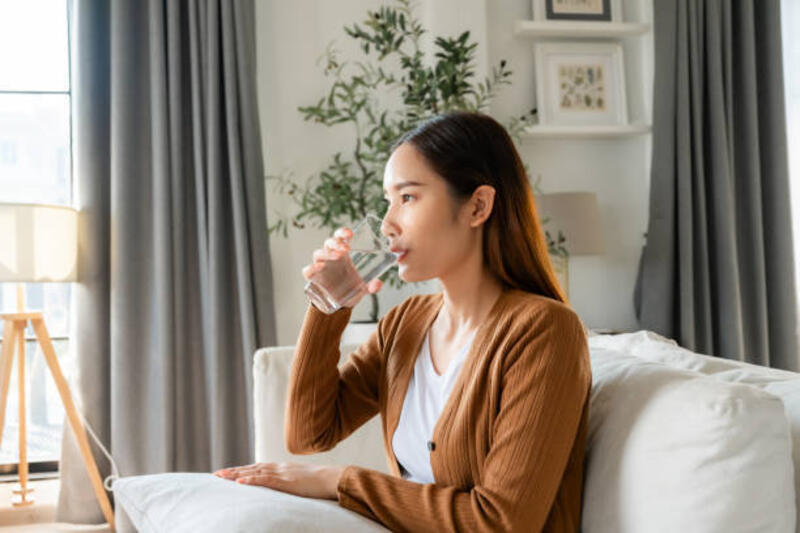 happy young asian woman drinking water while sitting on the sofa in the living room - wanita  minum air putih potret stok, foto, &amp; gambar bebas royalti